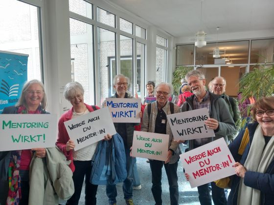 Eine Gruppe älterer Menschen hält Schilder hoch, auf denen "Mentoring wirkt" und "werde auch Du Mentor:in" steht.