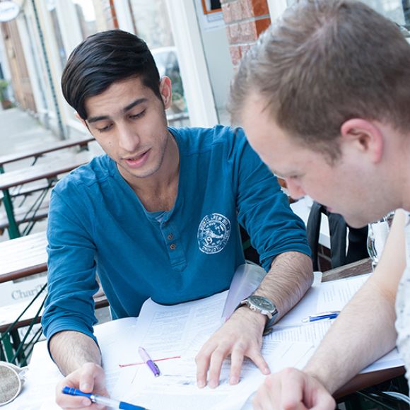 Ein Mentor und sein Mentee sitzen draußen an einem Tisch und blicken in verschiedene Unterlagen